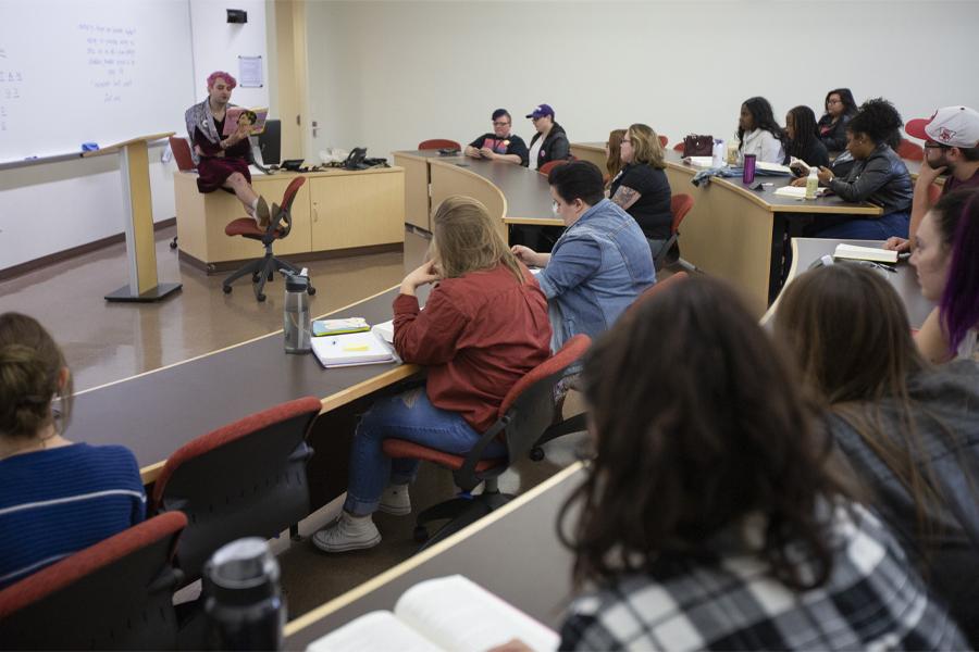 A person with pink hair reads from a book at the front of class.